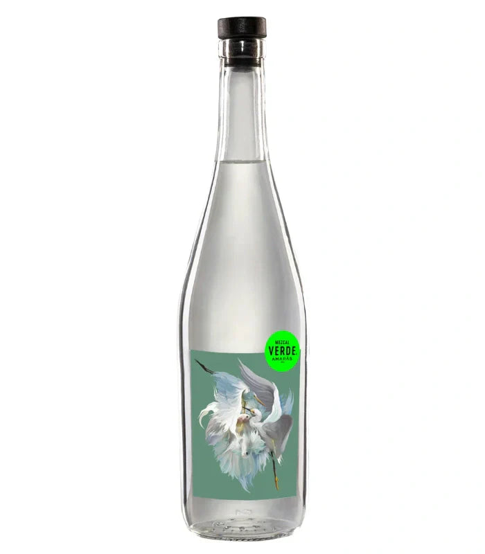 A clear glass bottle with a black cap, filled with clear liquid. The label shows a white flower on green and a small green circle reading MEZCAL VERDE. Product: Amaras Mezcal Verde Artist Series 2024 by Amaras.