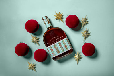 A Woodford Reserve Holiday Edition Bourbon 2025 “Designed by Markarian” 3 Pack is displayed amid red ornaments and gold star decorations, all set on a light blue background.